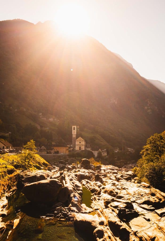 De ochtendzon komt boven de bergen uit in de Verzascavallei en verlicht een kerktoren en de rotsen in de rivier.