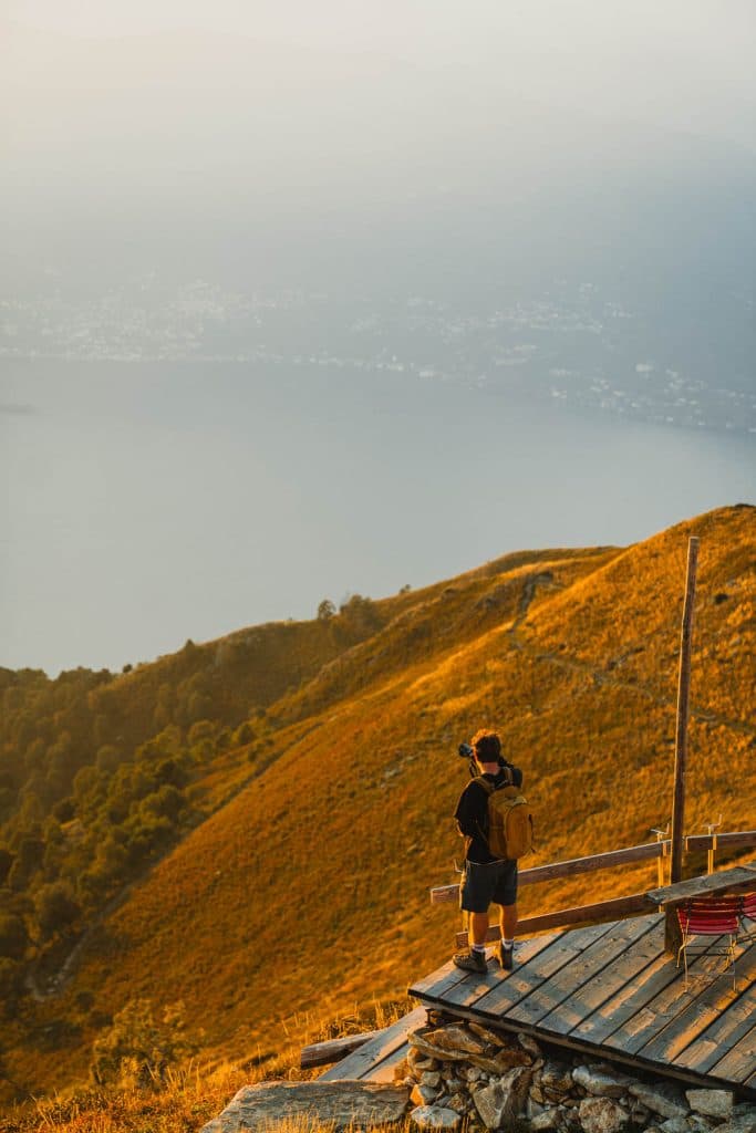 Een wandelaar staat op een houten terras op de Monte Gambarogno en fotografeert het Lago Maggiore in het avondlicht, boven de goudgele helling.