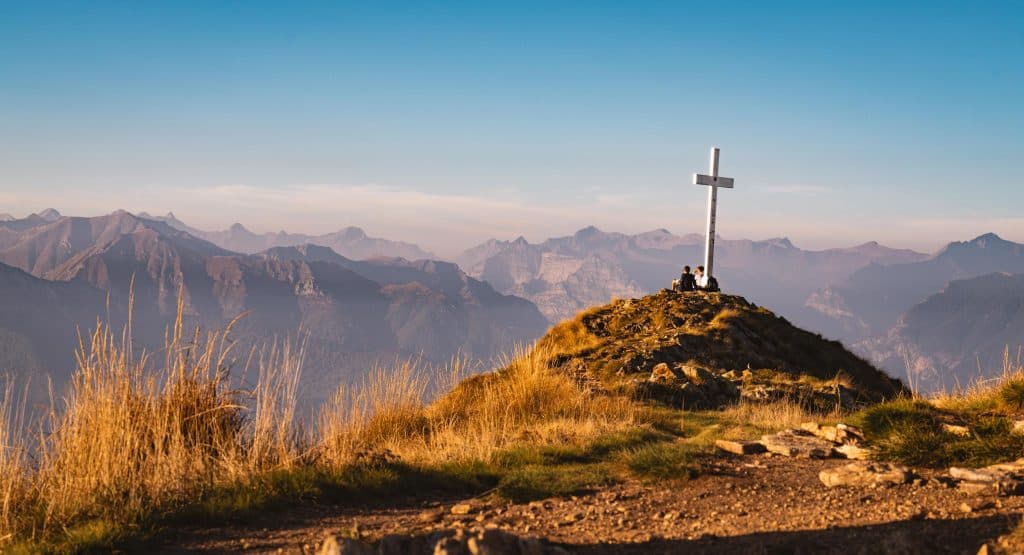 Het kruis op de top van de Monte Gambarogno tussen goudgeel gras, met een rij Alpentoppen in de avondzon.