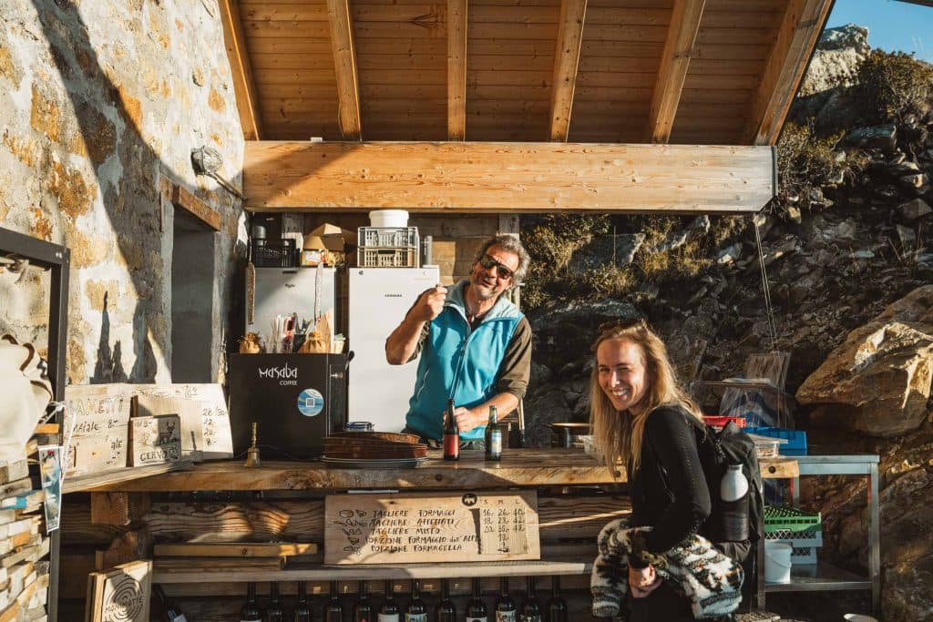 Een lachende man achter de houten bar van de berghut Capanna Gambarögn, met een vrouw met een rugzak die ernaast staat.