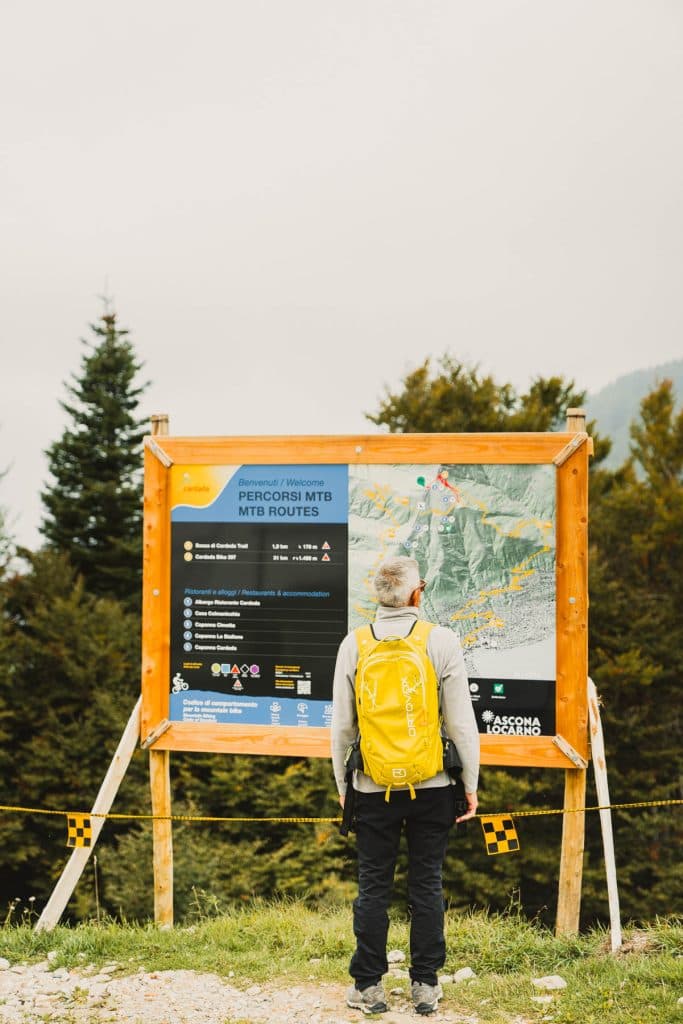 Een vrouw met een gele rugzak staat voor een groot bord met de mountainbikeroutes van Cardada en Cimetta.