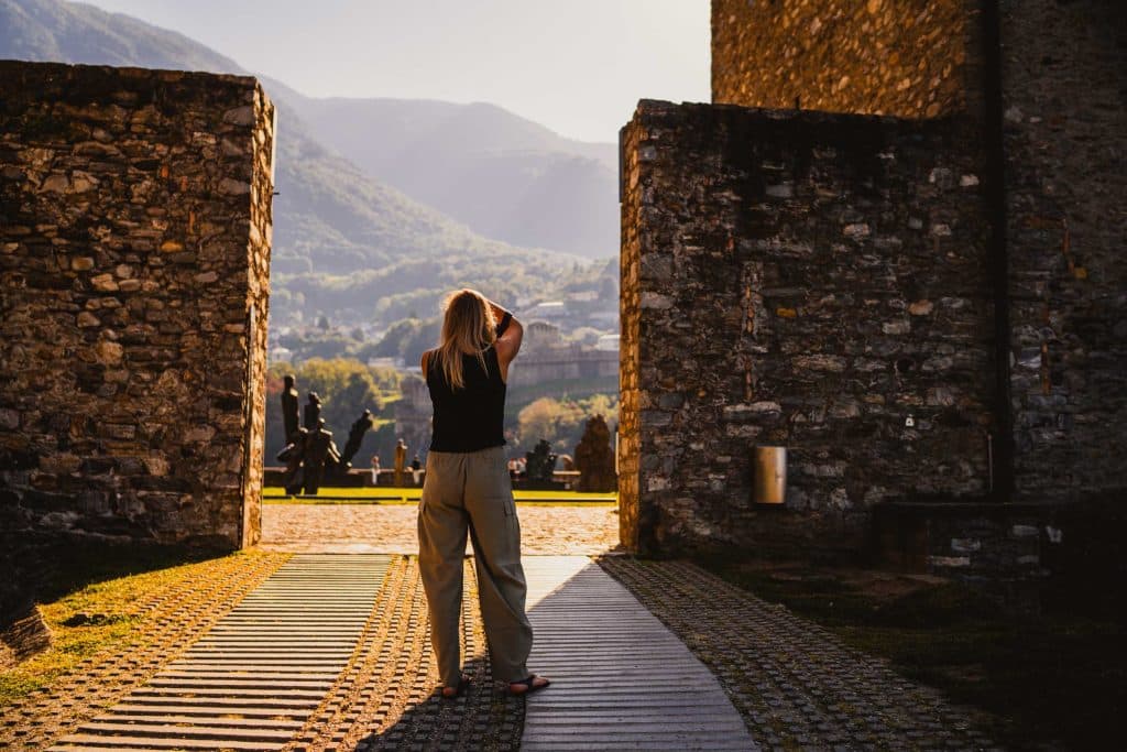 Een vrouw fotografeert tussen twee stenen muren op de binnenplaats van Castelgrande in Bellinzona, met de bergen erachter.