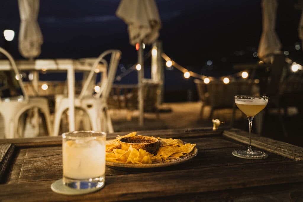 Een cocktail, een glas met citroen en een bord nacho's op een houten tafel, onder lichtsnoeren op een terras in Ascona.